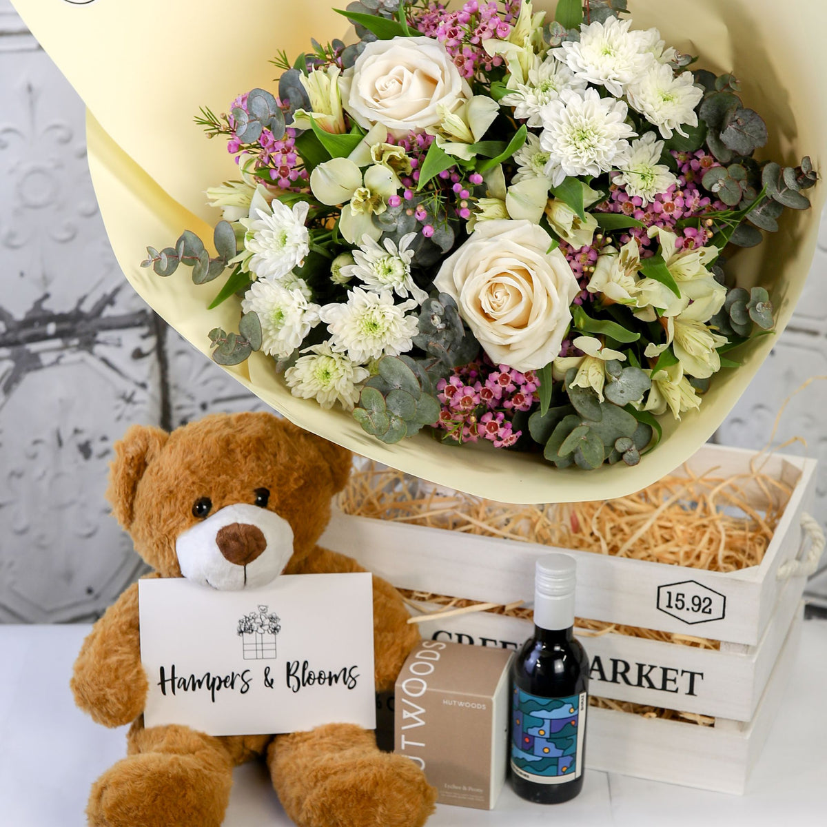 White and green bouquet above a white crate with a brown teddy holding a card, candle box and small bottle in front